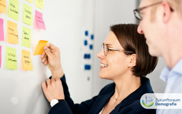 Eine Frau und ein Mann kleben Post-Its aus ein Whiteboard.